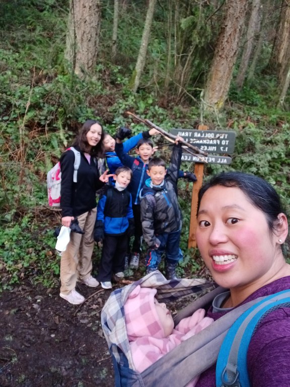 A mom takes a selfie with a sleeping baby and five kids and a trail sign in the background.