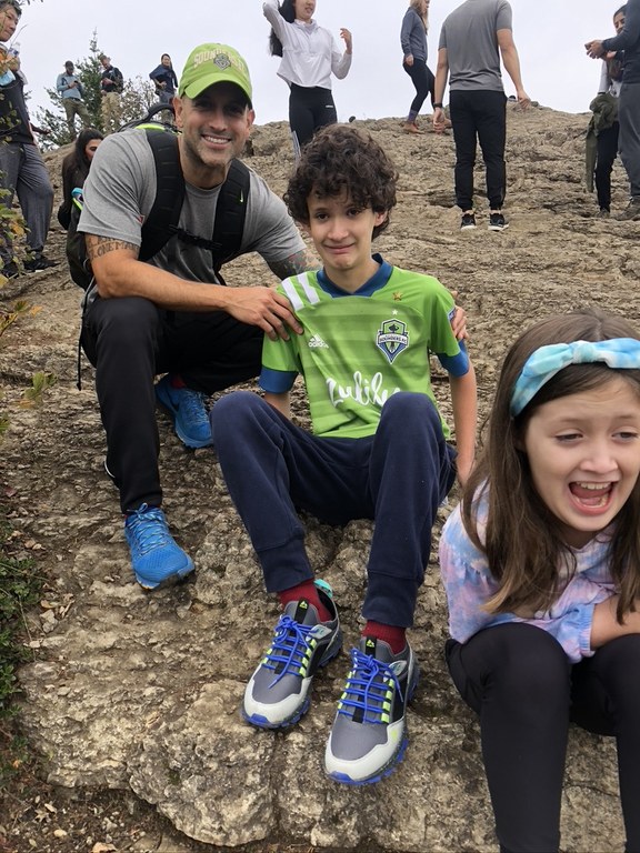 An adult and two kids pose for a photo on a large rock slab. The kids look dubious about the height. A number of hikers are visible in the background. 