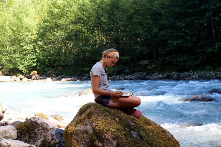 Reading by the river. Photo by McKenzie Carlson. A woman sits on a rock next to a river and reads a book. Photo by McKenzie Carlson.