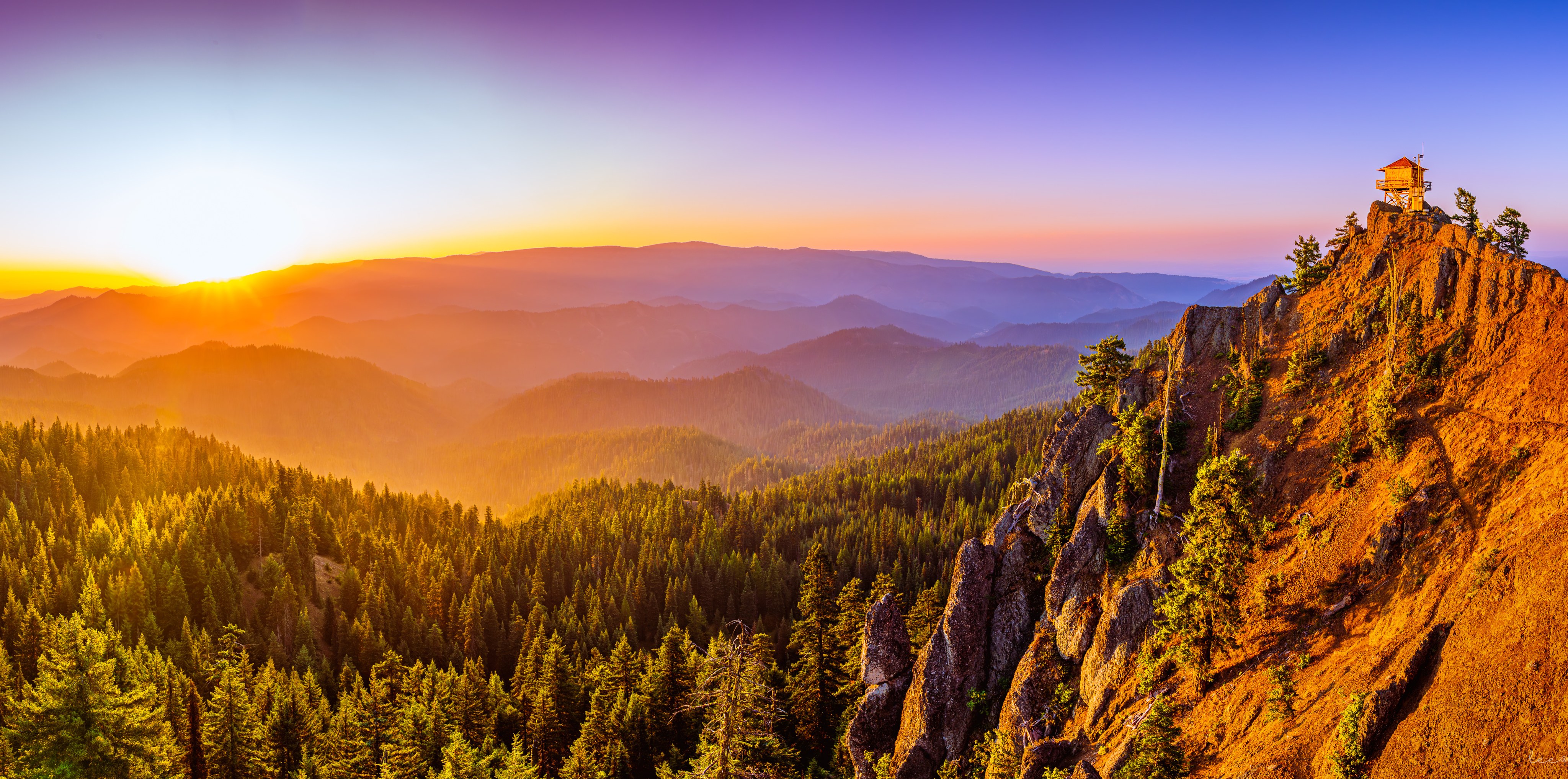 Historic fire lookout sits on a peak above a forested valley, glowing golden with the sun