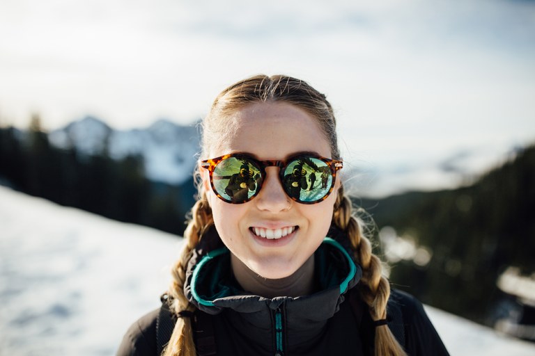 A woman smiles at the camera. The photographer is reflected in her sunglasses.