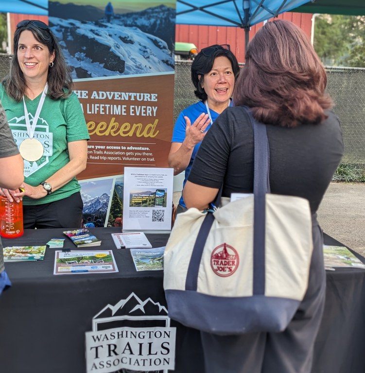 Outreach at Refuge Festival 2023 WTA representatives stand behind an outreach table at an outdoor event, engaged in conversation with attendees.