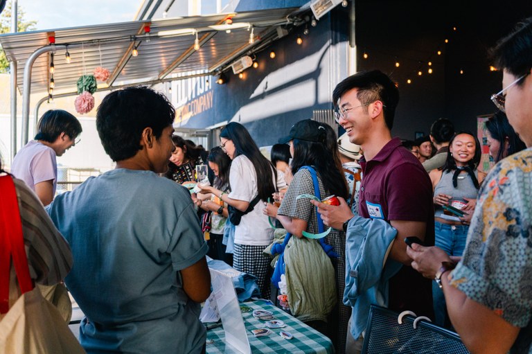 Outdoor Asian outreach event A group of people gathers around a table at an outdoor event, engaging happily. String lights and a corrugated roof create a festive atmosphere.