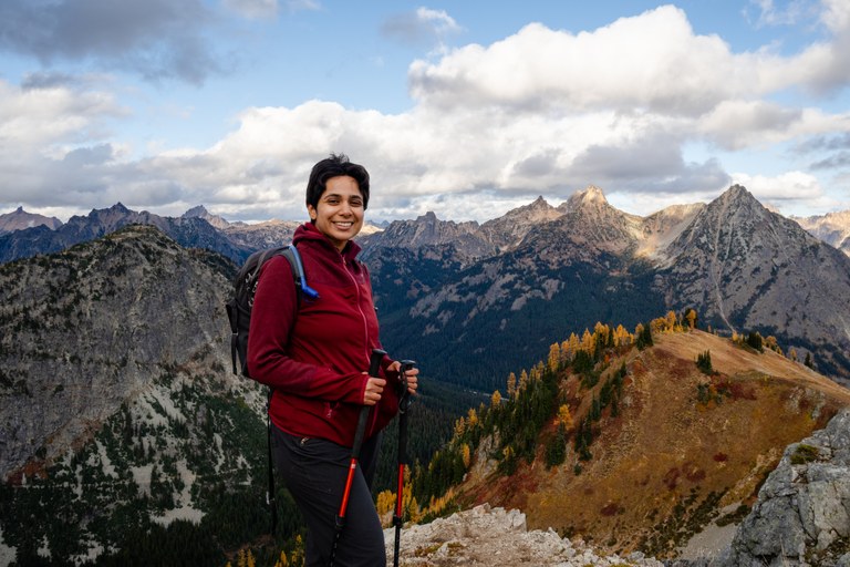 Saira Shahid portrait A person in a red jacket smiles on a mountain peak with trekking poles. Behind are rugged mountains and a partly cloudy sky, conveying adventure.