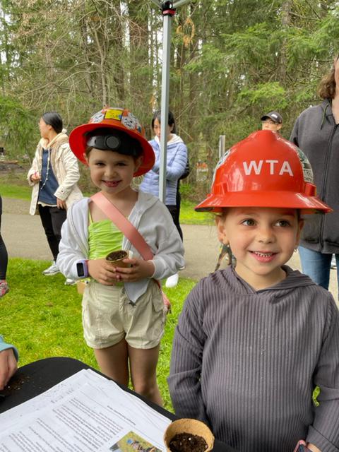 Two kids stand outside around a WTA booth outside. They are wearing orange WTA hardhats.
