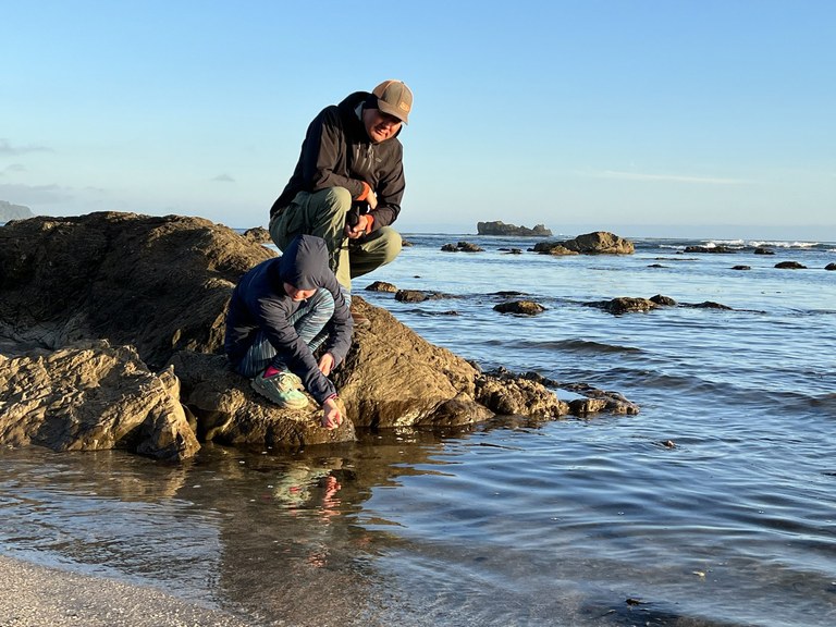 A man and kid sit on a rock and look in the saltwater.