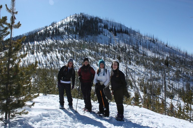Four women stand on snowshoes in a field full of packed snow with a snow-covered mountain behind them. 