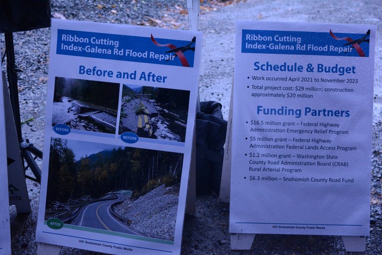 Signs show before and after of a road washed out, as well as budget numbers for the repairs required for the fix.