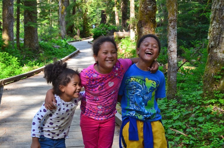 Three kids smile with their arms around each other, while standing on a boardwalk. 