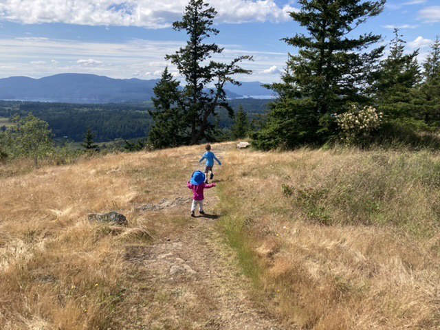 Two kids run on a trail in a grassy field with a wide view.