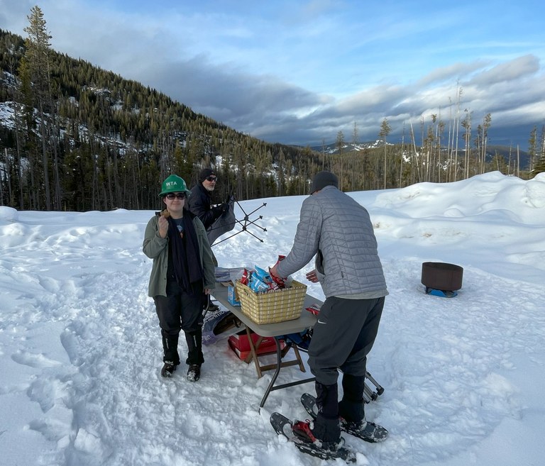 A person stands facing the camera holding a snack while standing next to a table of food set up in a snowy field