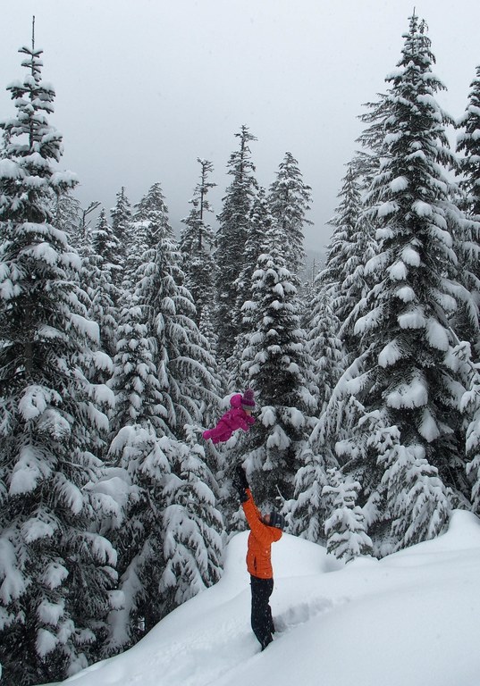 A person in the snow in a bright yellow jacket tosses a baby wearing bright pink high in the air.