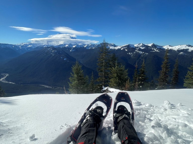 Snowshoes on a hiker's feet on the snow with mountains in the background. Photo by Birb.