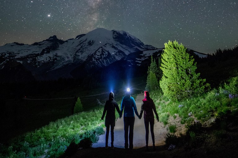 Three hikers hold hands while lookingup at a starry sky with their headlamps on. Mount Rainier is visible on the horizon and some green trees frame the photo. 