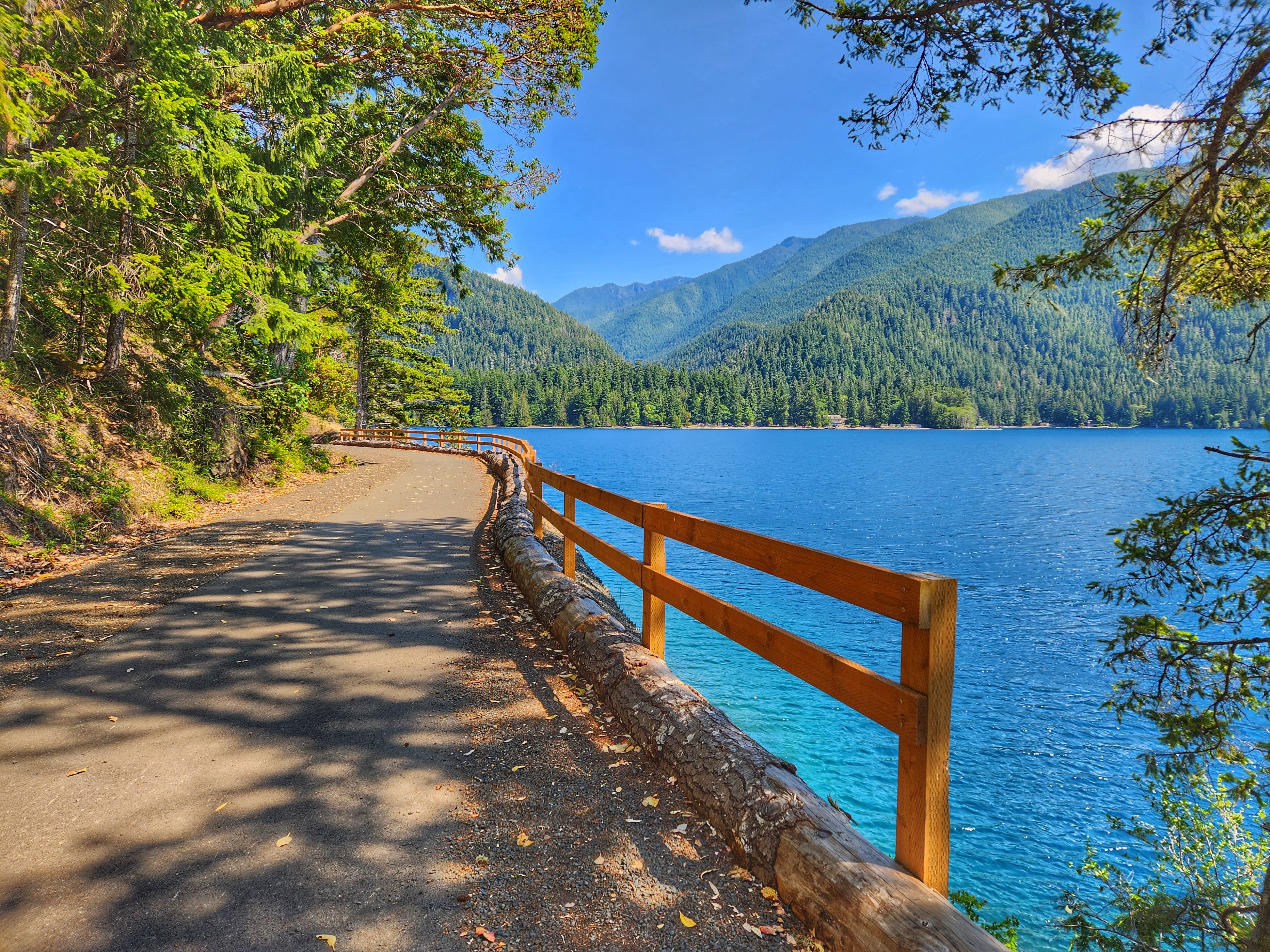 A wide, flat trail hugs the side of a blue lake on a sunny day
