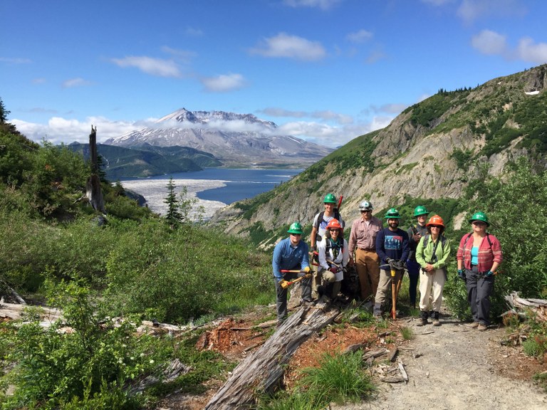 Group photo of a trail work crew at the Mount St. Helens National Monument with the volcano in the background on a sunny day. Photo by WTA staff.