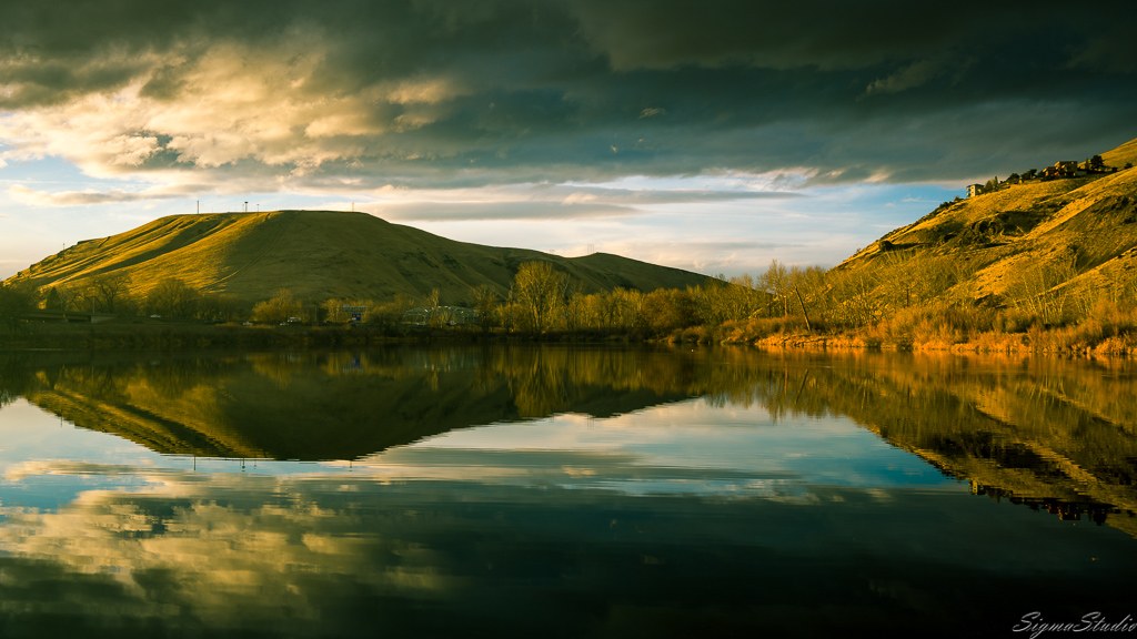 Brown hills and clouds are mirrored in the calm waters of a wide river