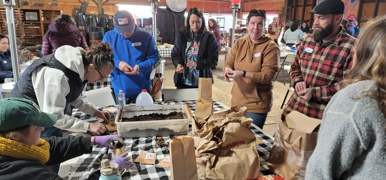 A group of adults gathers around a table in a workshop setting to plant seeds in small containers, with supplies and dirt bins visible on a black-and-white checkered tablecloth.