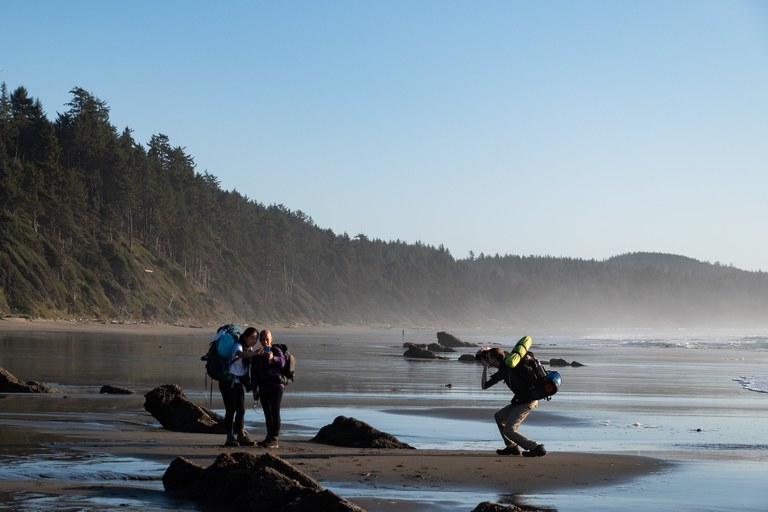 Man with a backpack takes a photo of two hikers taking a selfie on the beach. Photo by Mitchell Amundsen. 