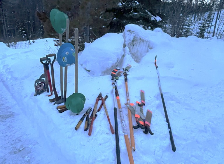 Several tools and a few hard hats are set up in the snow as if a crew is about to select them to use. 