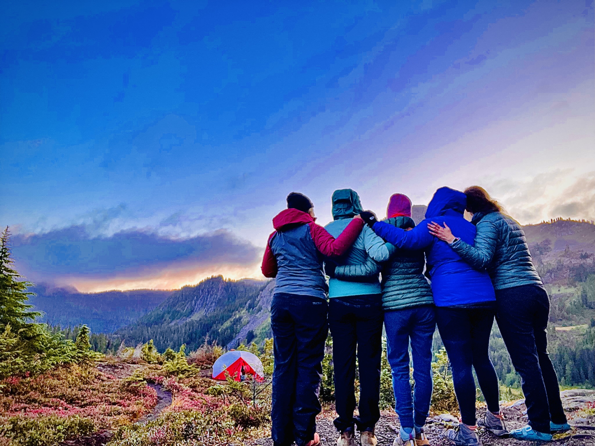 Five hikers stand with their arms around each other, overlooking a valley with a trail and tent nearby