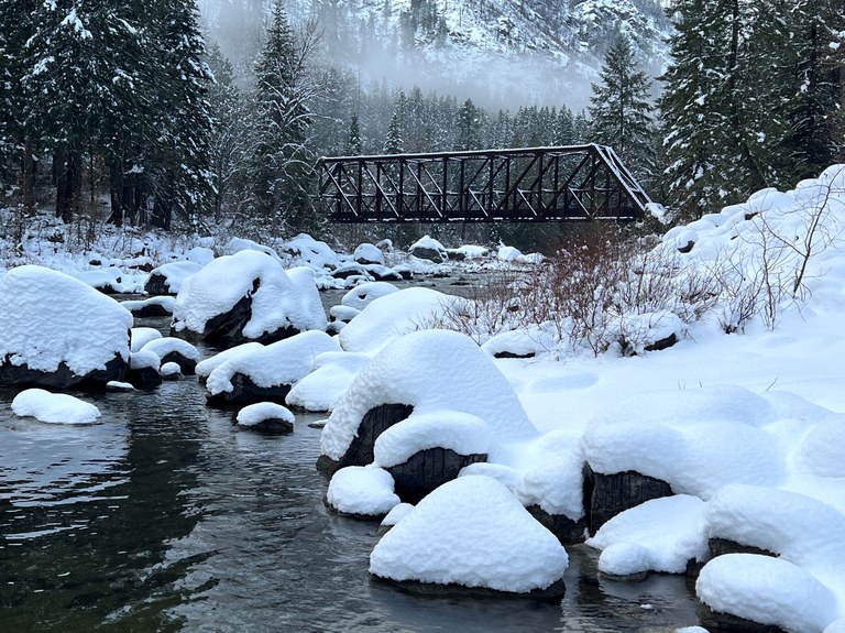 A snowy landscape along the river with a steel bridge and mountains in the background on the Tumwater Pipeline Trail. Photo by trip reporter joyoflandscapes.