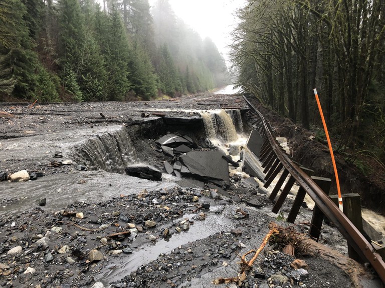 Water and rock debris covers U.S. 2 and the edge of the pavement crumbles off a slope next to a roadway barrier.