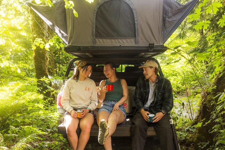 There's nothing like kicking back with friends after a long day of hiking. Photo by Carson Artec. Three young hikers enjoy beverages in tin camp cups on the tailgate of a vehicle with a popup tent above. Photo by Carson Artec.