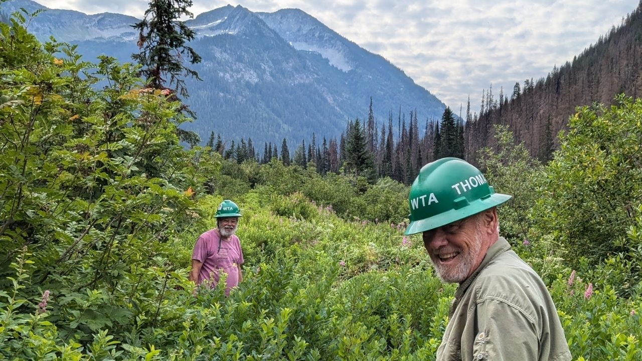 Two people in green hats smile at camera on trail covered in brush. Blue skies with clouds and mountains in the background.