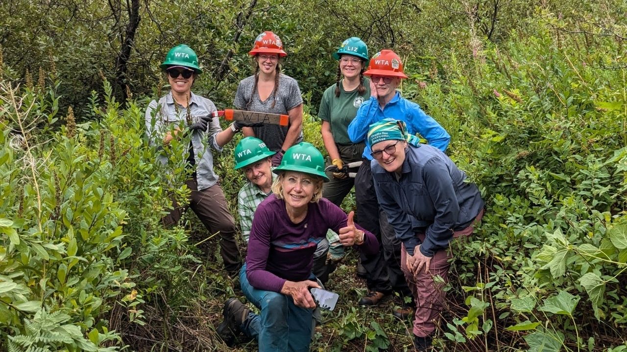 Seven people smile at camera. Some wearing orange and green hard hats.
