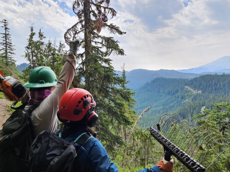 Two people face away from the camera and overlook a lush green valley. One has his hand triumphanly in the air.  
