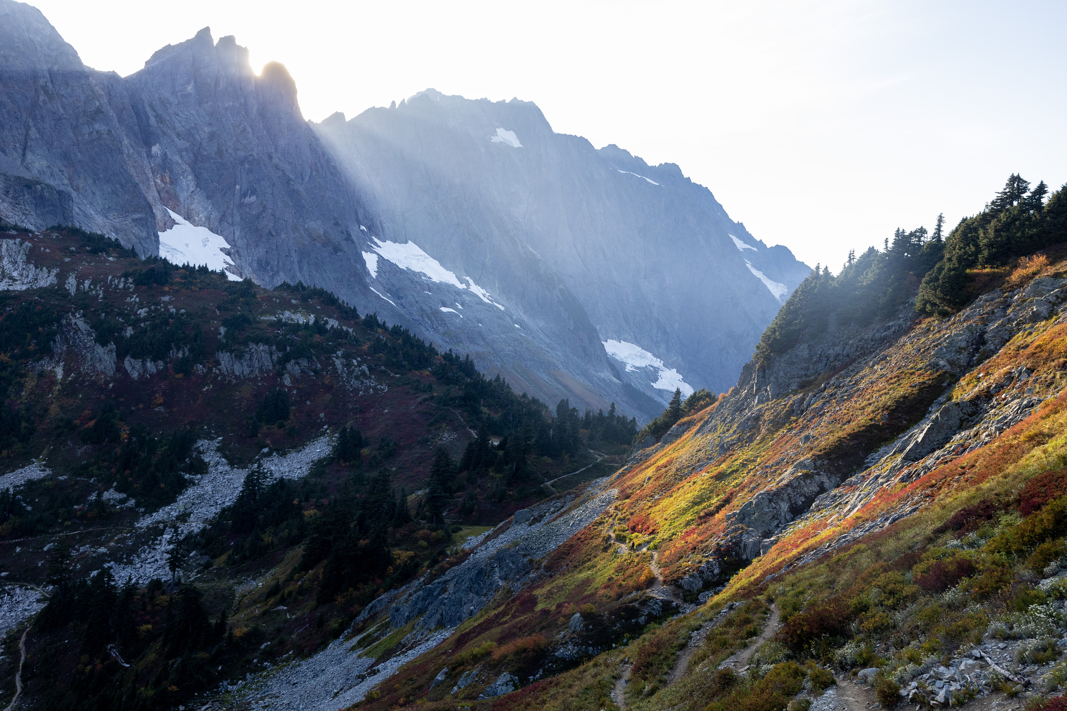 Sunbeams shine through a light wildfire smoke haze in North Cascades National Park as a hiker makes her way through Cascade Pass at the end of a fall day. Photo by Nathan Pilling
