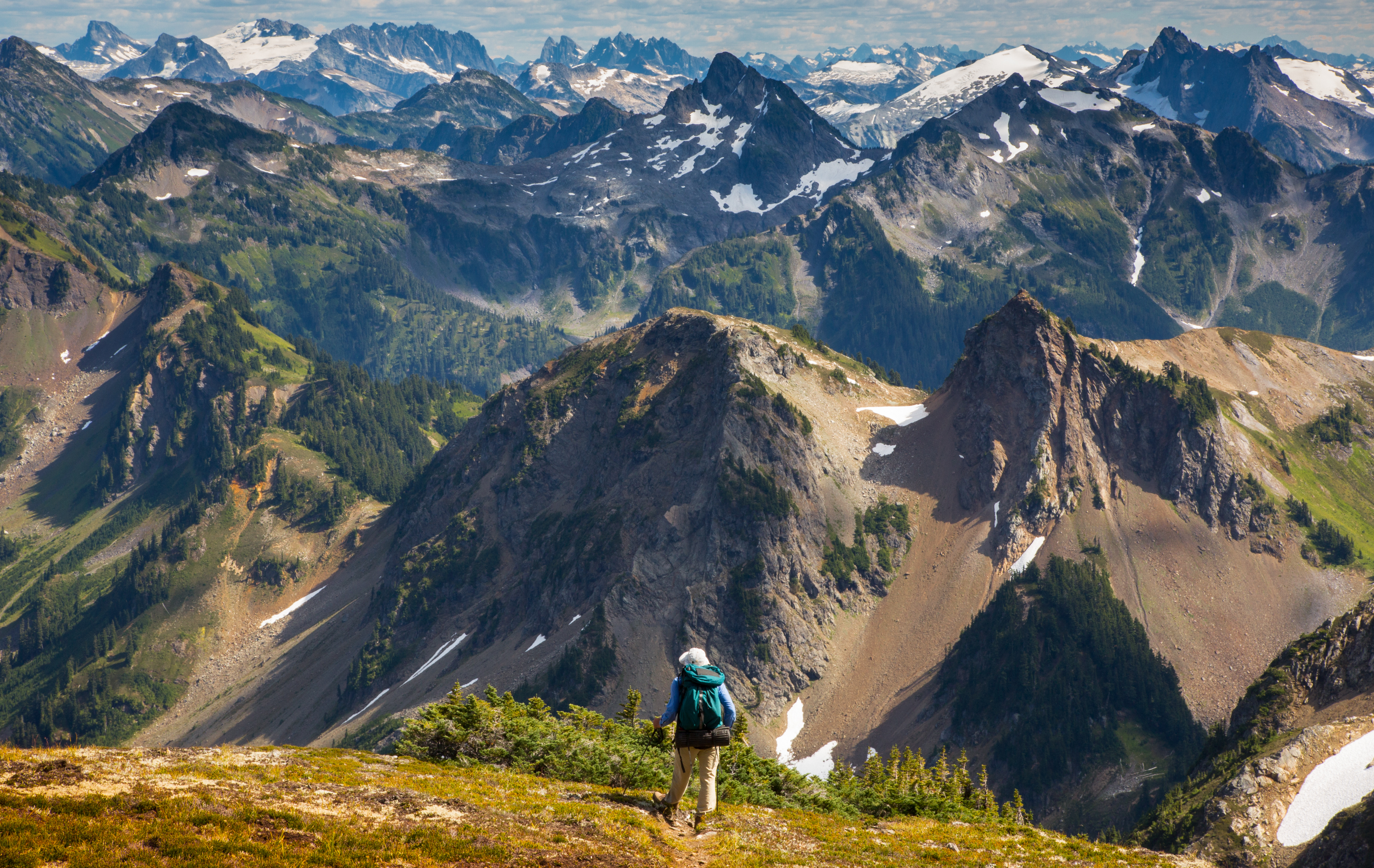 Image displays a hiker looking off at the mountain line, where there are layers of mountain peaks in front of him.