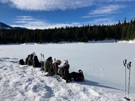 A group of youth sit down to eat lunch at the edge of a frozen lake, with trees and a blue sky in the background. 