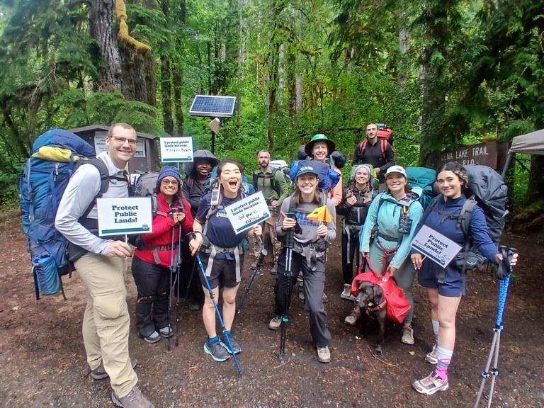 A dozen hikers stand at a trailhead, smiling and holding signs that say "Protect Public Lands"