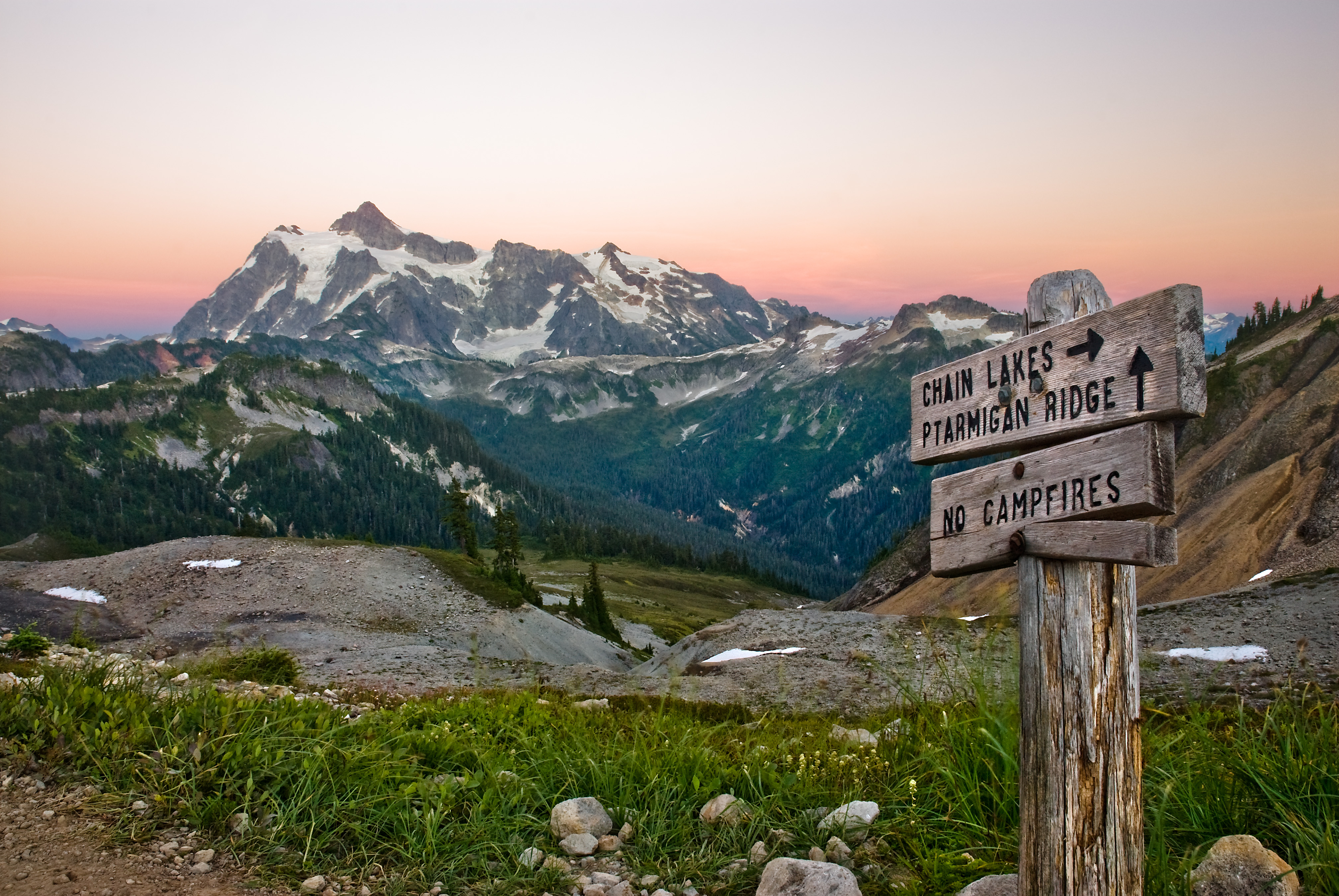 Wooden post labeling trails to Chain Lakes and Ptarmigan Ridge sits in front of rugged peaks and a glowing orange sky