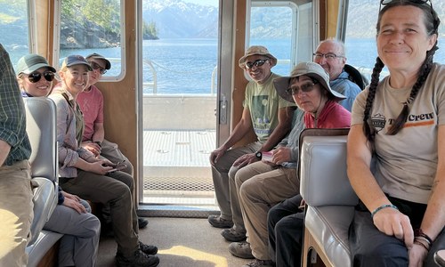 A small crew of people sit on a bus and sit facing the camera, smiling. A lake is visible out the back of the bus. 