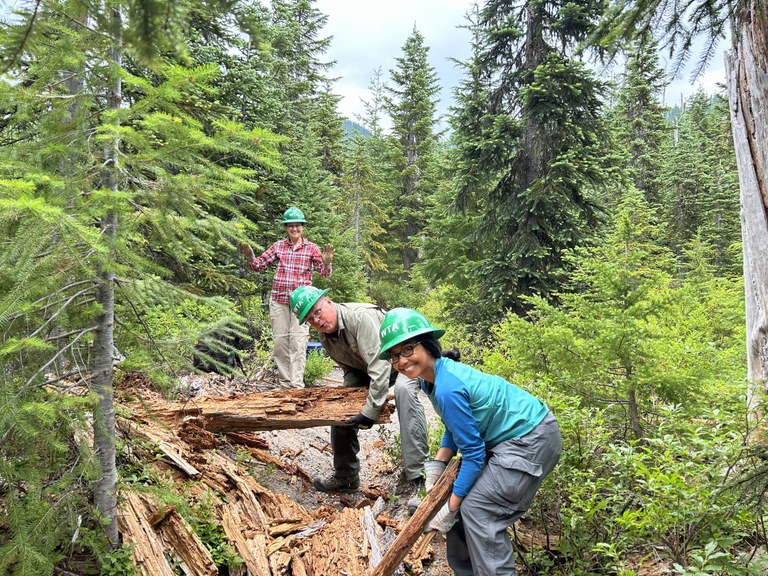 Three crew members stand and smile as they remove a large log from the trail at MSHI. 
